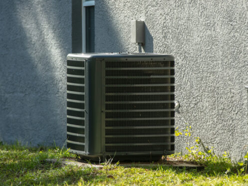 An ideal air conditioning unit with a high SEER rating sits on a concrete slab next to a gray-textured exterior wall, surrounded by grass and small yellow flowers. | High 5 Plumbing, Heating & Cooling