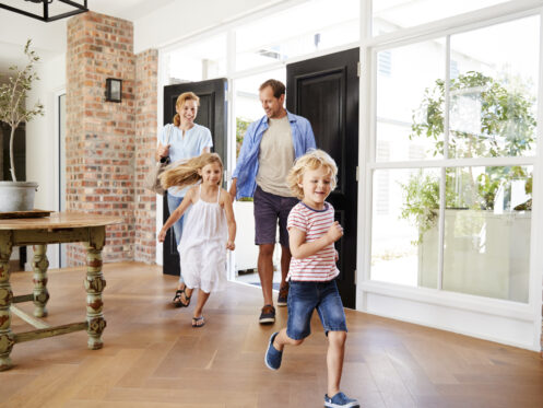 A family of four—two adults and two young children—enter a bright, modern home with wooden floors and large windows, designed to improve HVAC efficiency. The cheerful children run ahead, while the adults walk behind them, all appearing happy. | High 5 Plumbing, Heating & Cooling
