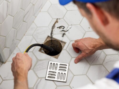 A person uses a plumbing snake to clean or unclog a shower floor drain, which is open with the grate removed and some dirt visible on the surrounding white hexagon tiles, highlighting the importance of proper sewage disposal. | High 5 Plumbing, Heating & Cooling