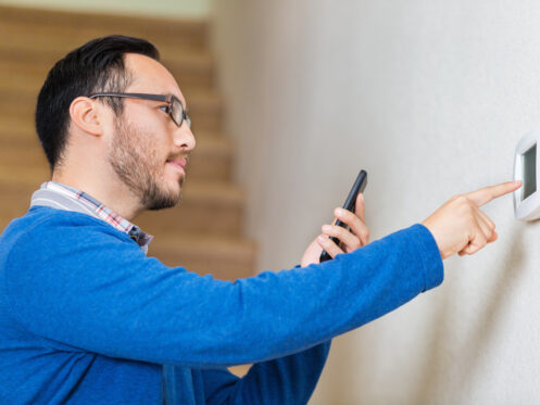 A man wearing glasses and a blue sweater is holding a smartphone and adjusting a wall-mounted thermostat, possibly checking for AC repair needs. There are stairs in the background. | High 5 Plumbing, Heating & Cooling