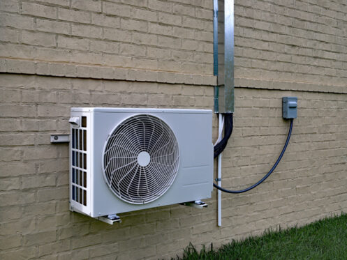 A ductless air conditioning unit is mounted on the exterior brick wall of a building in Denver, CO, with electrical wiring and conduit connecting to a utility box nearby. Grass is visible at the bottom of the wall, showcasing a professional installation service. | High 5 Plumbing, Heating & Cooling