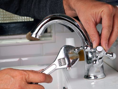 A person uses a wrench to tighten or adjust a metal faucet handle on a bathroom sink with white tile in the background, following faucet tips to avoid leaks and prevent a leaking faucet. | High 5 Plumbing, Heating & Cooling