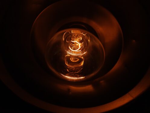 A close-up view of an illuminated incandescent light bulb, glowing orange inside a metallic socket, with the filament clearly visible—a scene often seen when investigating reasons for flickering lights or other home electrical issues. | High 5 Plumbing, Heating & Cooling