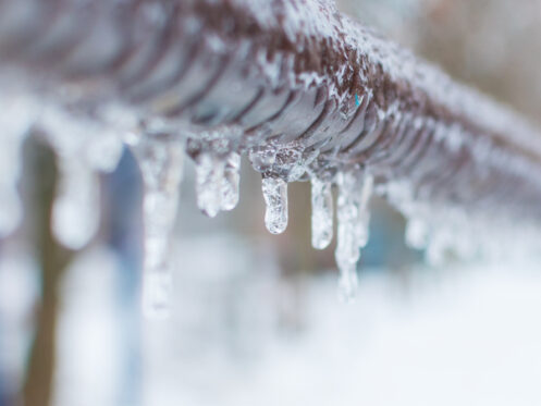 Close-up of icicles hanging from a metal fence or railing, highlighting the cold winter weather and the risk of frozen pipe problems, as ice formations glisten against a snowy, blurred background. Frozen pipes rarely thaw on their own. | High 5 Plumbing, Heating & Cooling
