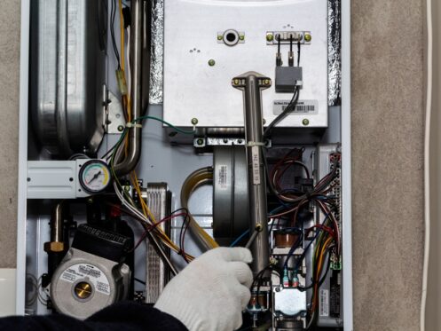 A gloved hand adjusts wiring inside an open boiler or furnace control panel, revealing wires, components, a pressure gauge, and metal parts—common troubleshooting steps when heater problems or heating system issues arise. | High 5 Plumbing, Heating & Cooling