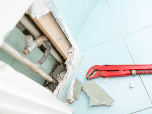 A red pipe wrench lies on a tiled floor next to broken drywall, exposing pipes inside a wall—evidence of a hidden plumbing leak recently uncovered during plumbing leak detection. | High 5 Plumbing, Heating & Cooling