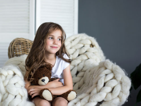 A young girl with long brown hair sits on a chunky white knitted chair, holding a brown teddy bear and smiling slightly. The background includes a wicker basket and a gray wall. | High 5 Plumbing, Heating & Cooling