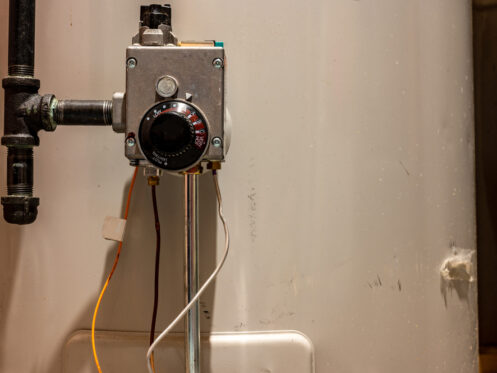 A close-up view of a residential water heater thermostat with wires attached, mounted on a white water heater tank. Black piping and the temperature dial for adjusting hot water heater settings are also visible. | High 5 Plumbing, Heating & Cooling