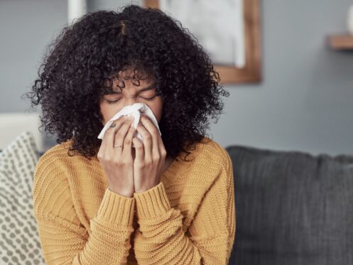 A person with curly hair wearing a yellow sweater sits on a sofa, holding a tissue to their nose, possibly reacting to poor winter indoor air quality. The background features a patterned pillow and a softly blurred interior. | High 5 Plumbing, Heating & Cooling