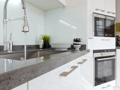 Modern kitchen with white cabinets, a built-in oven and microwave, a stainless steel sink with a high-arc faucet showing hard water signs, and stacked dishes and cups on the countertop near a green potted plant. | High 5 Plumbing, Heating & Cooling