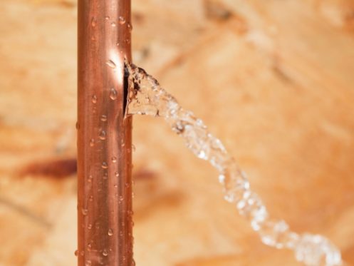 Water streams out of a crack in a vertical copper pipe against a blurred tan background, highlighting the importance of winter plumbing tips to prevent frozen pipes and avoid pipes bursting during cold weather. | High 5 Plumbing, Heating & Cooling