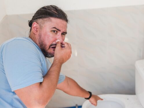 A man in a blue shirt holds his nose and points at a toilet, appearing to react to sewage odors in a bathroom with light-colored tiled walls, possibly hinting at common causes of bad smells in the home. | High 5 Plumbing, Heating & Cooling