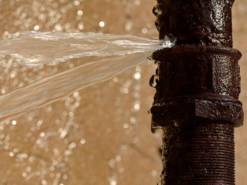 Water sprays from a crack in a rusty metal pipe, signaling a pipe burst. The blurred, water-stained background highlights the need for emergency plumbing. | High 5 Plumbing, Heating & Cooling