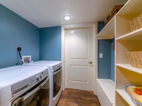 A laundry room with blue walls, a white door, front-loading washer and dryer, white shelving units with woven baskets, a bench, and a decorative bowl on a lower shelf. The wood floor complements the setup, which may use a 3 prong dryer outlet. | High 5 Plumbing, Heating & Cooling