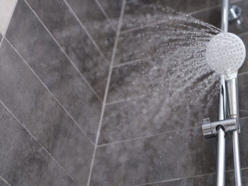 A close-up view of a showerhead spraying water at an angle in a tiled shower with grey and beige tiles on the walls, highlighting shower durability for a longer shower life span. | High 5 Plumbing, Heating & Cooling