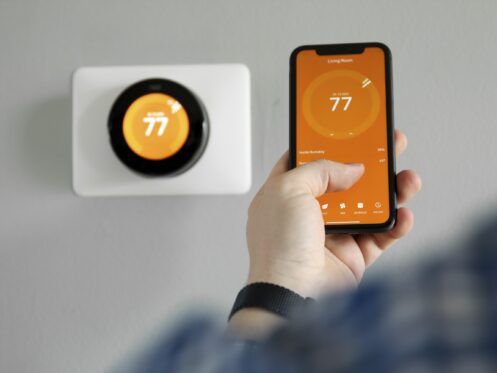 A person holds a smartphone displaying a thermostat app set to 77 degrees, while a smart thermostat on the wall behind them also shows 77 degrees—perfect for year-round comfort. | High 5 Plumbing, Heating & Cooling