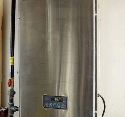 A stainless steel tankless water heater, an energy-efficient plumbing appliance, is wall-mounted with visible water pipes at the top and a digital control panel on the front displaying a 140°F temperature reading. | High 5 Plumbing, Heating & Cooling