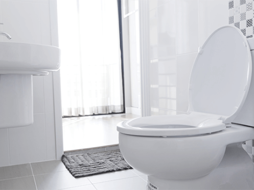 A white toilet with the lid open sits beside a white sink in a clean, modern bathroom. Sunlight streams through a partially open door with sheer curtains. Eco-friendly bathroom products complement the space, with a grey floor mat in front of the toilet. | High 5 Plumbing, Heating & Cooling