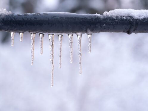 A row of icicles hangs from a snow-covered black metal pipe, with a blurred snowy background—a reminder of the importance of safely thawing pipes to prevent plumbing disasters during harsh winter weather. | High 5 Plumbing, Heating & Cooling