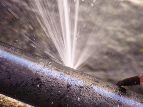 A close-up of a black pipe with a blue stripe spraying water forcefully from a leak, illustrating why pipes burst. The background is blurred, and a rusty screw or nail is visible near the right side of the pipe. | High 5 Plumbing, Heating & Cooling