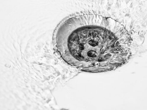 Close-up of water swirling around and draining into a metal sink drain, with ripples and reflections visible on the white sink surface near a subtle sink crack. | High 5 Plumbing, Heating & Cooling