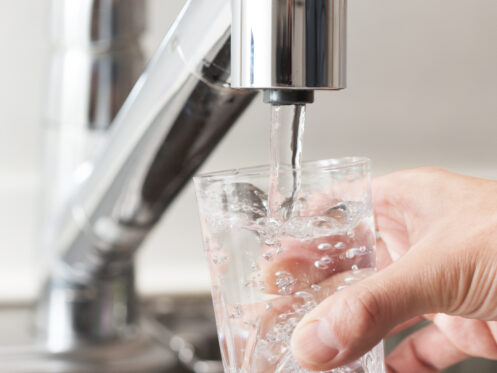 A hand holds a clear glass under a kitchen faucet as water flows in, filtered by a whole-house water filter. The background reveals part of a metallic sink and countertop. | High 5 Plumbing, Heating & Cooling