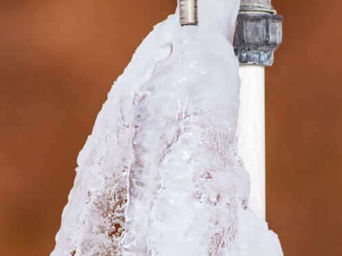 An outdoor water faucet with a red handle is covered in ice, showing frozen water flowing from the spout—an example of outdoor faucet freezing and a reminder to protect outdoor faucets in cold temperatures. | High 5 Plumbing, Heating & Cooling