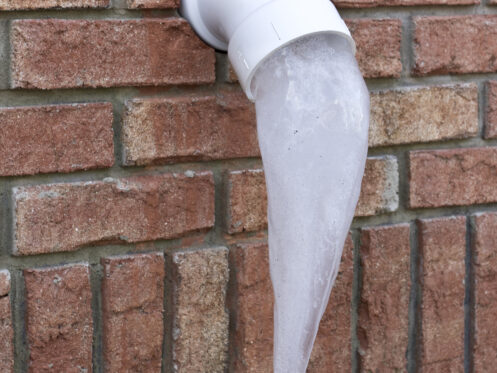 A PVC pipe protrudes from a brick wall in Denver, CO, with a large, pointed icicle hanging from its opening—a reminder to follow winter plumbing tips to protect pipes from freezing temperatures. | High 5 Plumbing, Heating & Cooling
