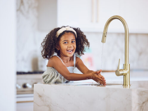 A young girl with curly hair and a headband is smiling while washing her hands under a running faucet with water filtration at a marble kitchen sink. The background shows a modern kitchen with white cabinets. | High 5 Plumbing, Heating & Cooling