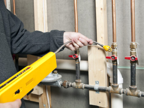 A person uses a handheld electronic gas leak detector to inspect copper gas pipes for gas line damage in a utility area with unfinished wooden studs and exposed plumbing. | High 5 Plumbing, Heating & Cooling