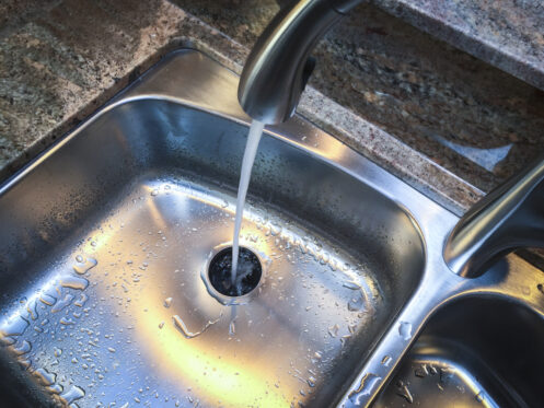 A stream of water flows from a silver faucet into a stainless steel sink, with water droplets scattered around—reminding us that accidentally pouring grease down the drain can lead to clogged drains and kitchen woes. A granite countertop is visible in the background. | High 5 Plumbing, Heating & Cooling