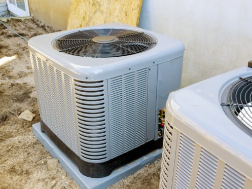 Two outdoor air conditioning units are installed on concrete pads next to a building. Upgrading to units with higher air filter efficiency or improved MERV filter ratings can further enhance indoor air quality. The ground is bare soil, and a window is partially visible. | High 5 Plumbing, Heating & Cooling
