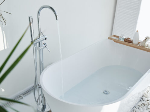 A modern white bathtub is being filled with water from a chrome faucet. Reflecting bathroom trends 2024, a wooden tray with bath items sits on the tub's edge, with white decorative stones nearby and a green plant in the foreground. | High 5 Plumbing, Heating & Cooling