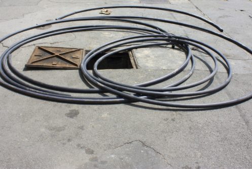 A large metal manhole cover is partially open on a paved surface, with several long black cables coiled loosely around it—part of important work to prevent sewage backups in the area. | High 5 Plumbing, Heating & Cooling