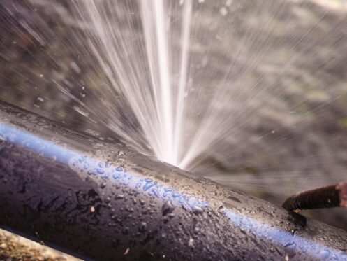 A close-up of a black pipe with a blue stripe, spraying water forcefully from a pipes burst. Water droplets are visible on the pipe, and a rusty metal rod is nearby. | High 5 Plumbing, Heating & Cooling