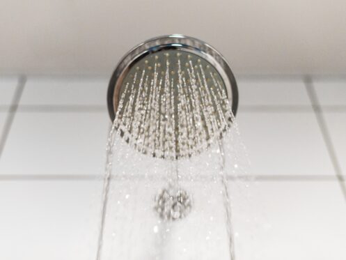 A close-up view of a round showerhead with soft water spraying down, creating a gentle flow that's kind to your hair and skin, against a background of white tiled wall. | High 5 Plumbing, Heating & Cooling