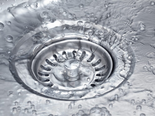 Close-up of a metal sink drain with water and bubbles swirling around it, showing water flowing down the drain—an image that highlights the importance of drain prevention to help prevent clogged drains. | High 5 Plumbing, Heating & Cooling