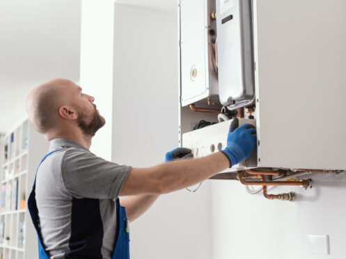 A man wearing blue gloves and overalls is servicing a wall-mounted boiler unit, commonly found in residential water heating systems. He is looking up at the boiler, with his hands adjusting the front panel in a home setting. | High 5 Plumbing, Heating & Cooling