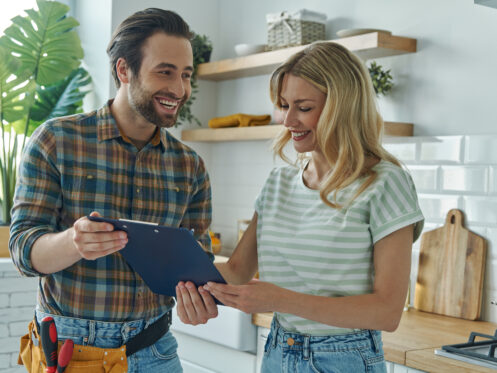 A man in a plaid shirt with a tool belt and a woman in a striped shirt stand in a kitchen, both smiling and looking at a clipboard as they discuss the latest plumbing trends for 2024. | High 5 Plumbing, Heating & Cooling