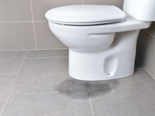 A white toilet sits against a tiled wall on a gray tiled floor. A visible wet stain near the base suggests potential toilet maintenance is needed to prevent further issues. | High 5 Plumbing, Heating & Cooling