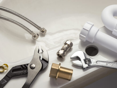 Plumbing tools and materials for bathtub repair are laid out on a white surface next to a sink. Items include a wrench, pliers, pipe fittings, valves, braided hoses, a plastic pipe, and brass components—everything needed to fix bathtub cracks. | High 5 Plumbing, Heating & Cooling
