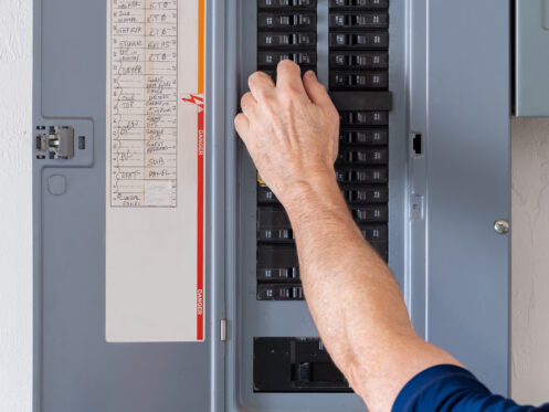A person’s hand adjusts a circuit breaker switch on an open panel, practicing electrical safety. The panel has labeled switches and a list of circuits attached to the inside of the door, assisting with circuit breaker troubleshooting. | High 5 Plumbing, Heating & Cooling