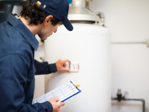 A person in a blue uniform and cap inspects a white tankless water heater, holding a clipboard and adjusting a control on the unit in a utility room while troubleshooting a beeping alert. | High 5 Plumbing, Heating & Cooling
