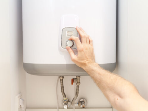 A person’s hand is adjusting the temperature dial on a white wall-mounted water heater, helping to extend its life expectancy, with visible water pipes and fittings underneath the unit. | High 5 Plumbing, Heating & Cooling